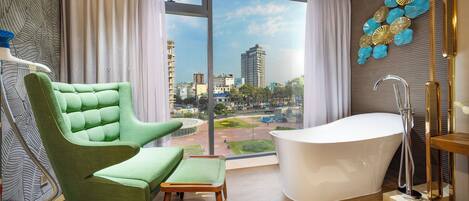 A modern bathroom with a freestanding bathtub, a green armchair, and a view of the cityscape through large windows.