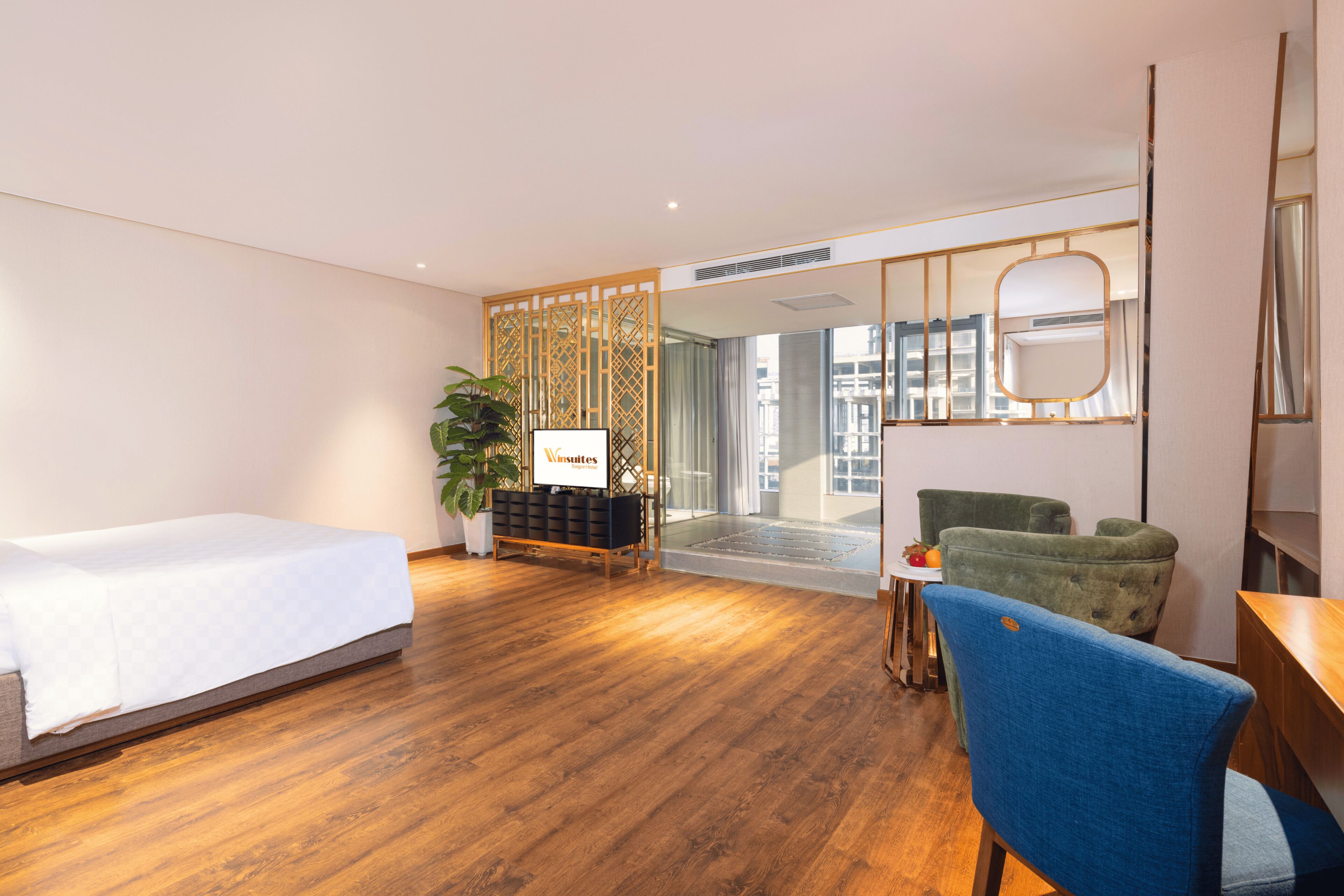 A hotel room with a wooden floor, a bed, a desk, a chair, a potted plant, and a view of the city through a glass door.