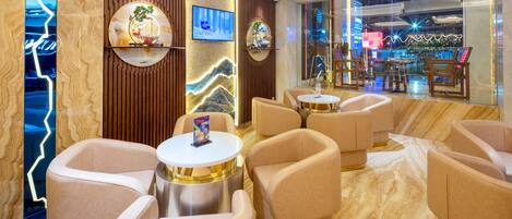 A modern hotel lobby with plush seating, a TV screen, and a large window offering a view of the cityscape.