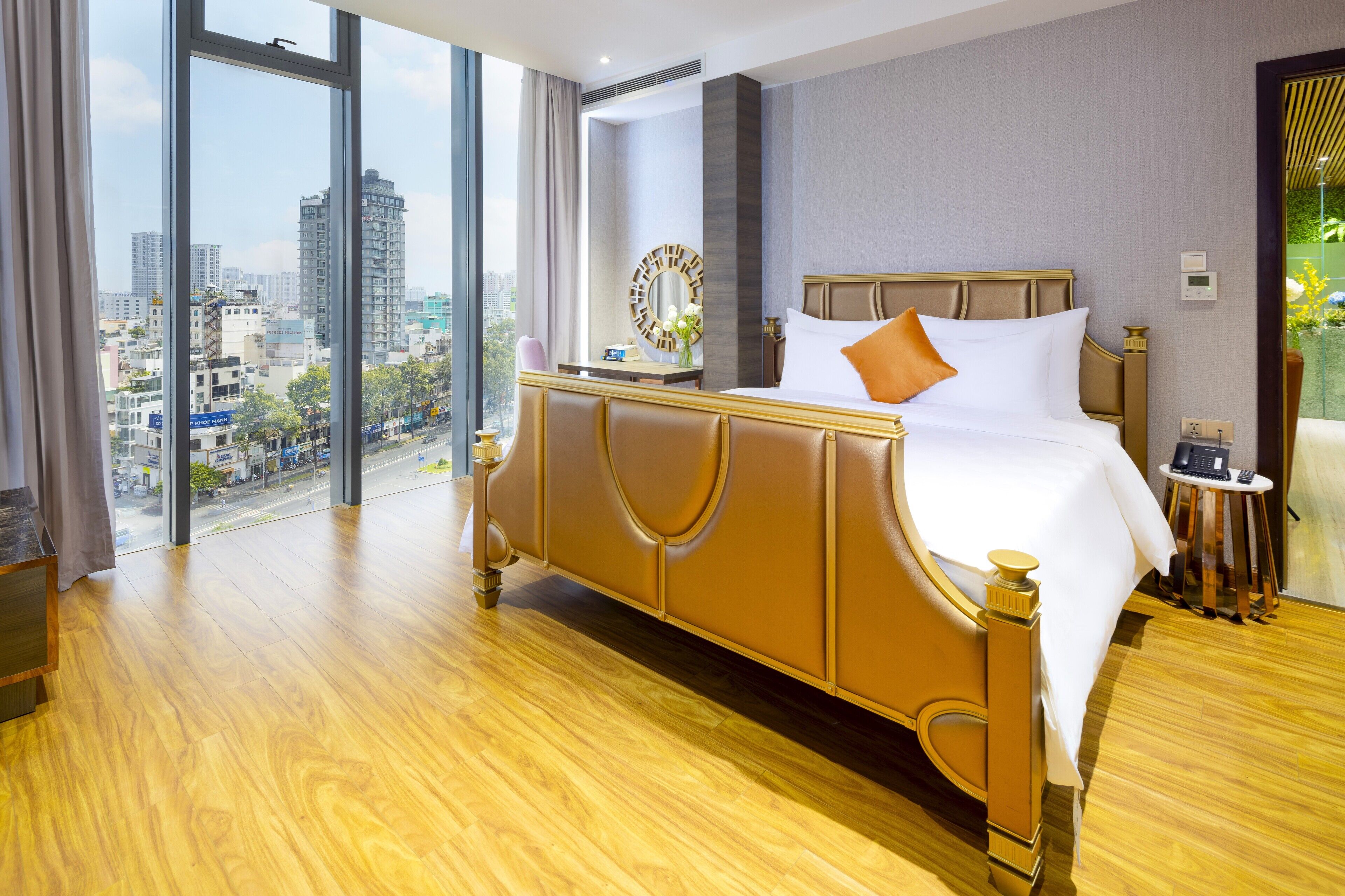 A hotel room with a large bed, a bedside table, a mirror, and a view of the city through large windows.