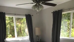 Un dormitorio con cama, escritorio, ventilador de techo y ventana con cortinas.