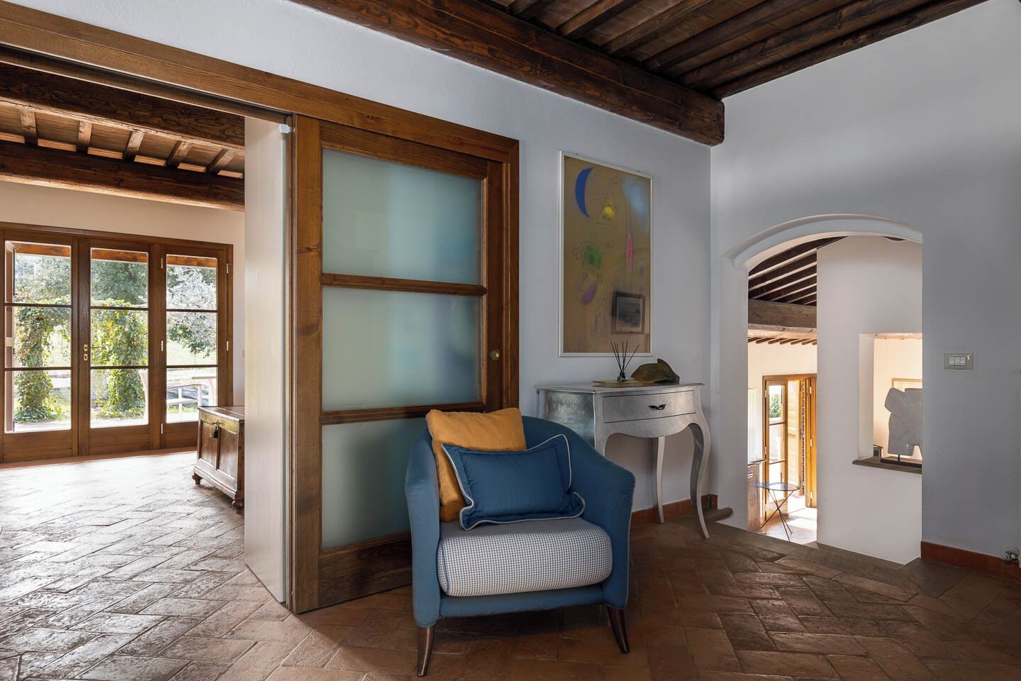 A room with a wooden ceiling, a blue armchair, a desk, and a door leading to a garden.