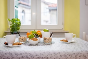 Disposição para o café da manhã, com toalha de mesa branca, duas xícaras brancas, copo de cereal, prato de biscoitos e tigela de frutas.