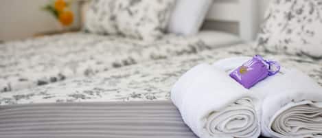 A neatly made bed with a floral-patterned bedspread and pillows, a folded white towel with a purple tag, and a small vase with a yellow flower on the nightstand.