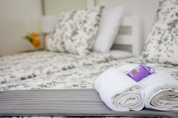 Cama bem arrumada com colcha e almofadas estampadas com flores, uma toalha branca dobrada com etiqueta roxa e um pequeno vaso com uma flor amarela na mesa de cabeceira.