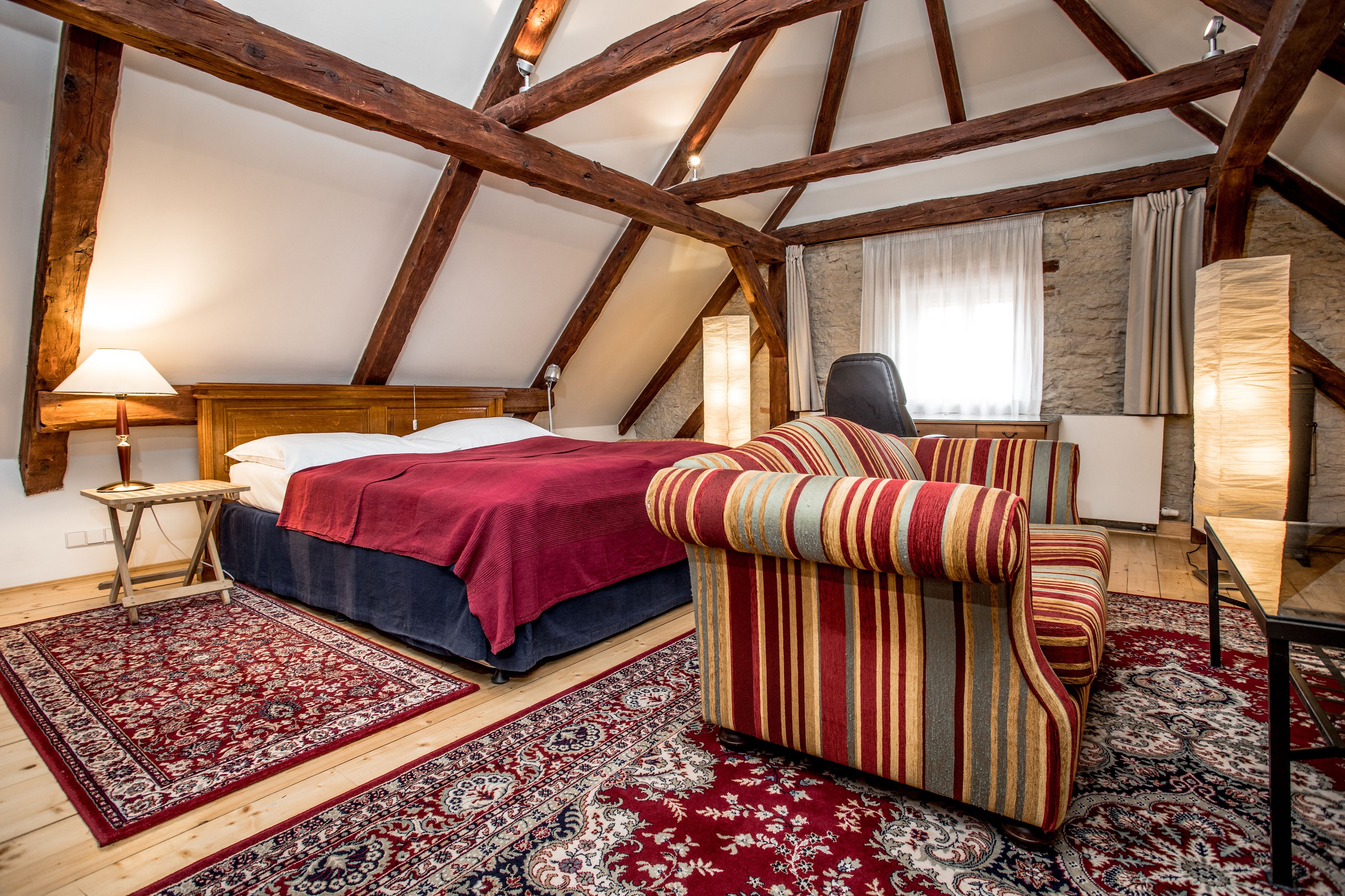 Un dormitorio con techo de madera, una cama con manta roja, un sillón a rayas y una pequeña mesa con lámpara.