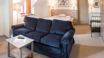 A hotel room with a dark blue sofa, a small table with a remote control and a bottle, a bed with a headboard, and a painting on the wall.