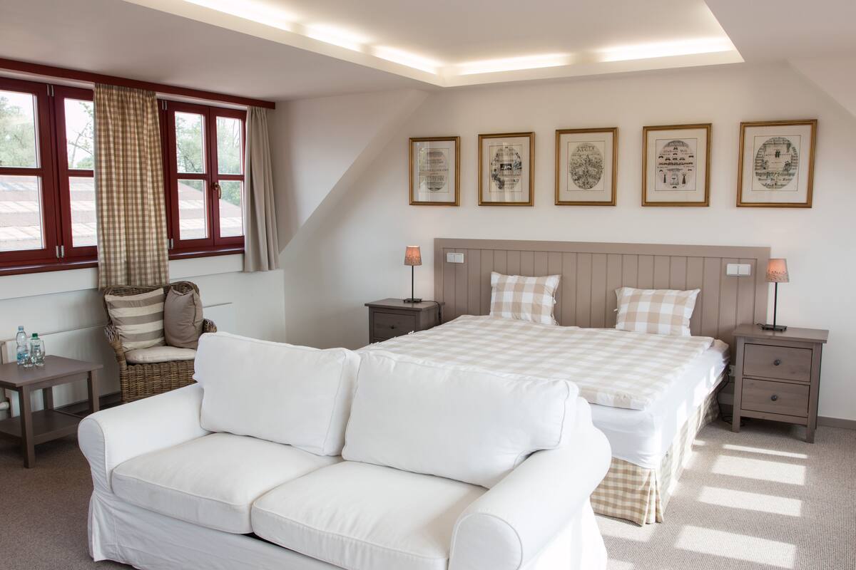A bedroom with a large bed, a white sofa, a wicker chair, and a nightstand with a lamp. There are four framed pictures on the wall.