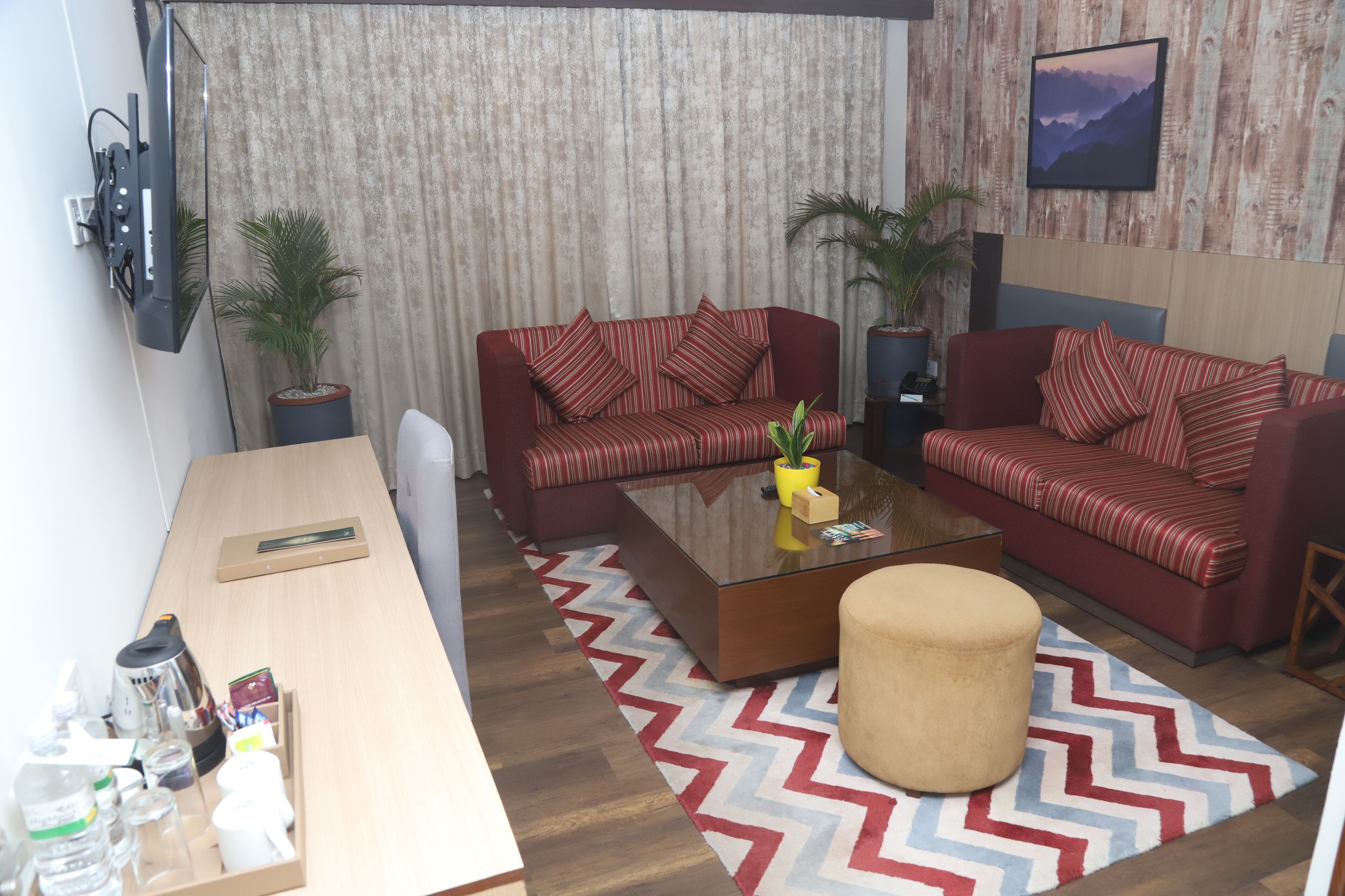 A modern living room with two red sofas, a glass coffee table, and a wooden desk with a chair. There are two potted plants, a patterned rug, and a framed picture on the wall.