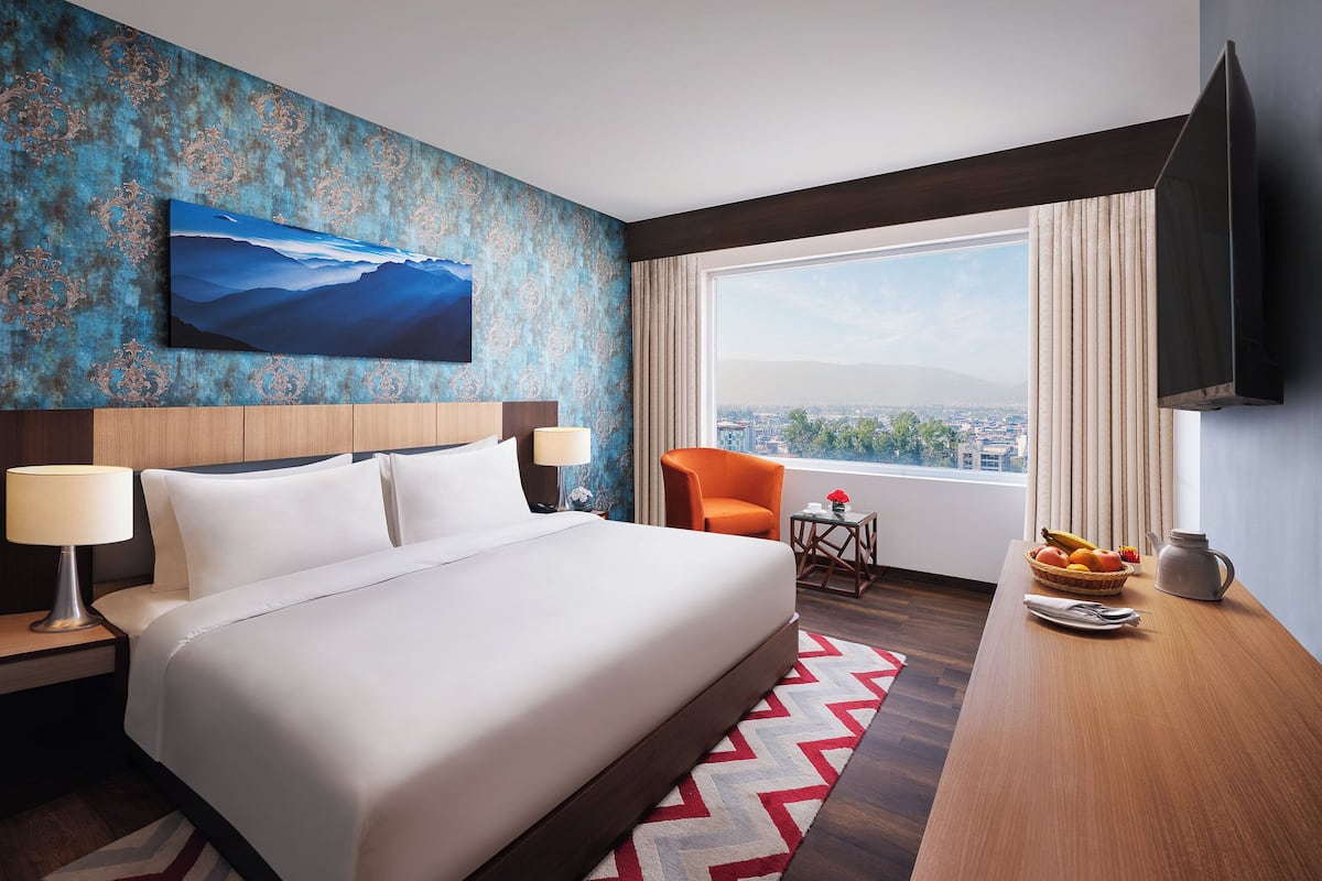 A hotel room with a large bed, a desk with a fruit bowl, a chair, a TV, and a city view.