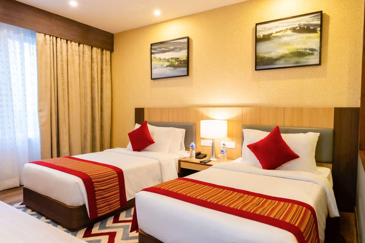 A hotel room with two beds, each with a red and gold striped bedspread, white pillows, and a wooden headboard. There are two paintings on the wall, a nightstand with a lamp, and a window with curtains.