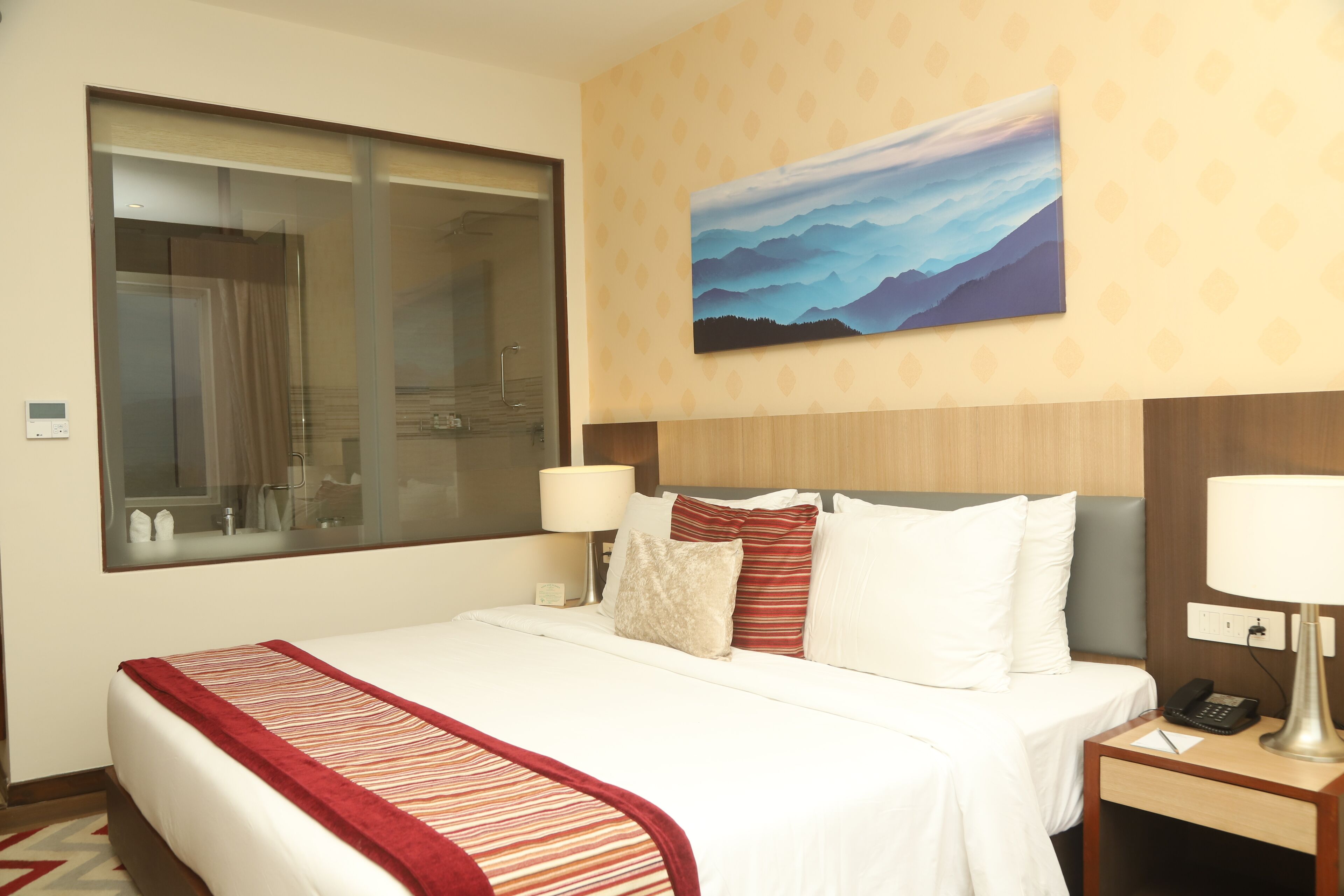 A hotel room with a large bed, a nightstand with a lamp, a painting on the wall, and a window with a view of the mountains.