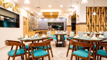 A modern dining area with wooden tables and teal chairs, a kitchen in the background, and decorative wall panels.