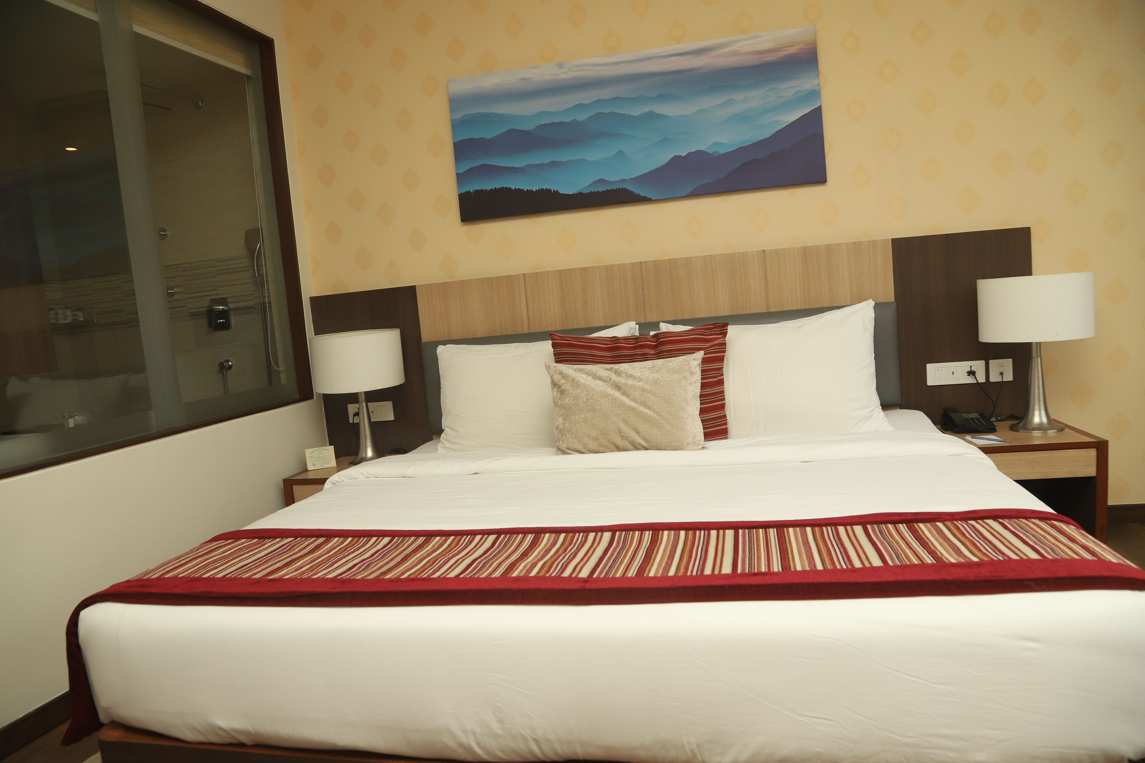 A hotel room with a large bed, two bedside tables with lamps, a painting on the wall, and a window with a view of mountains.
