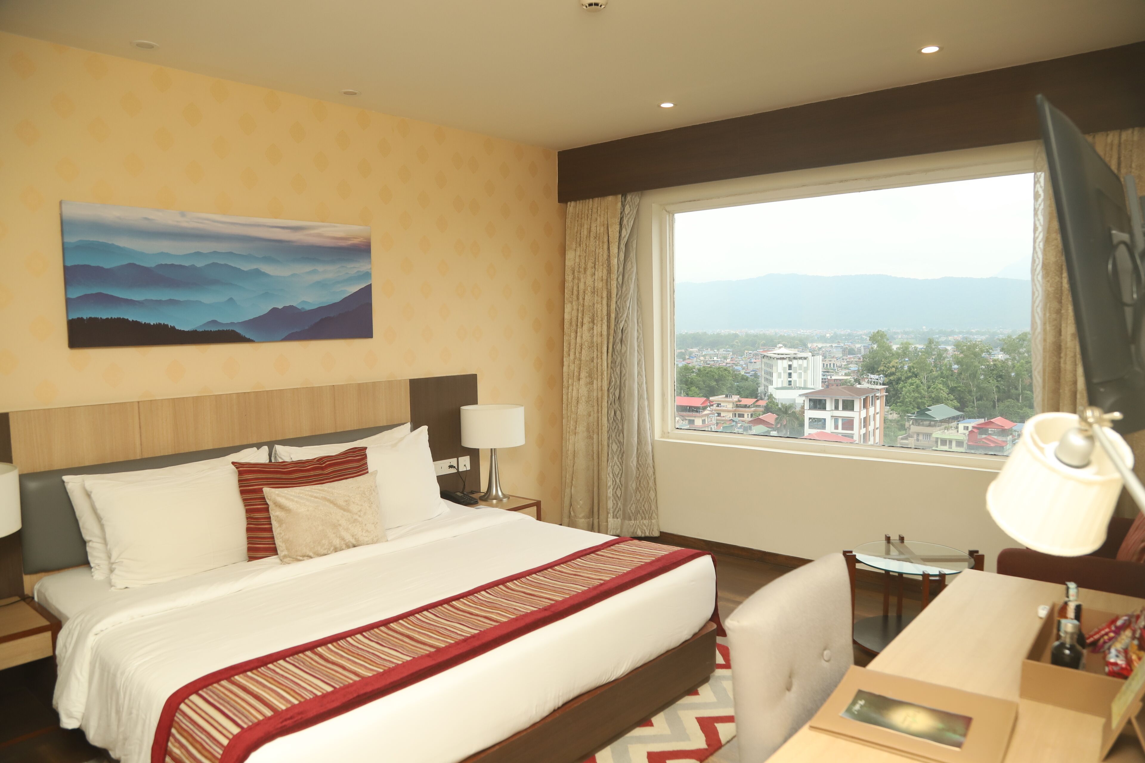 A hotel room with a large bed, a desk, a chair, a window with a view of buildings and mountains, and a painting on the wall.