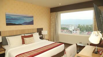 A hotel room with a large bed, a desk, a chair, a window with a view of buildings and mountains, and a painting on the wall.