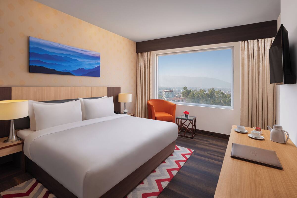 A hotel room with a large bed, a desk with a laptop, a chair, a small table with a vase, and a painting on the wall.