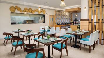 A restaurant with wooden tables and chairs, a kitchen in the background, and hanging pendant lights.
