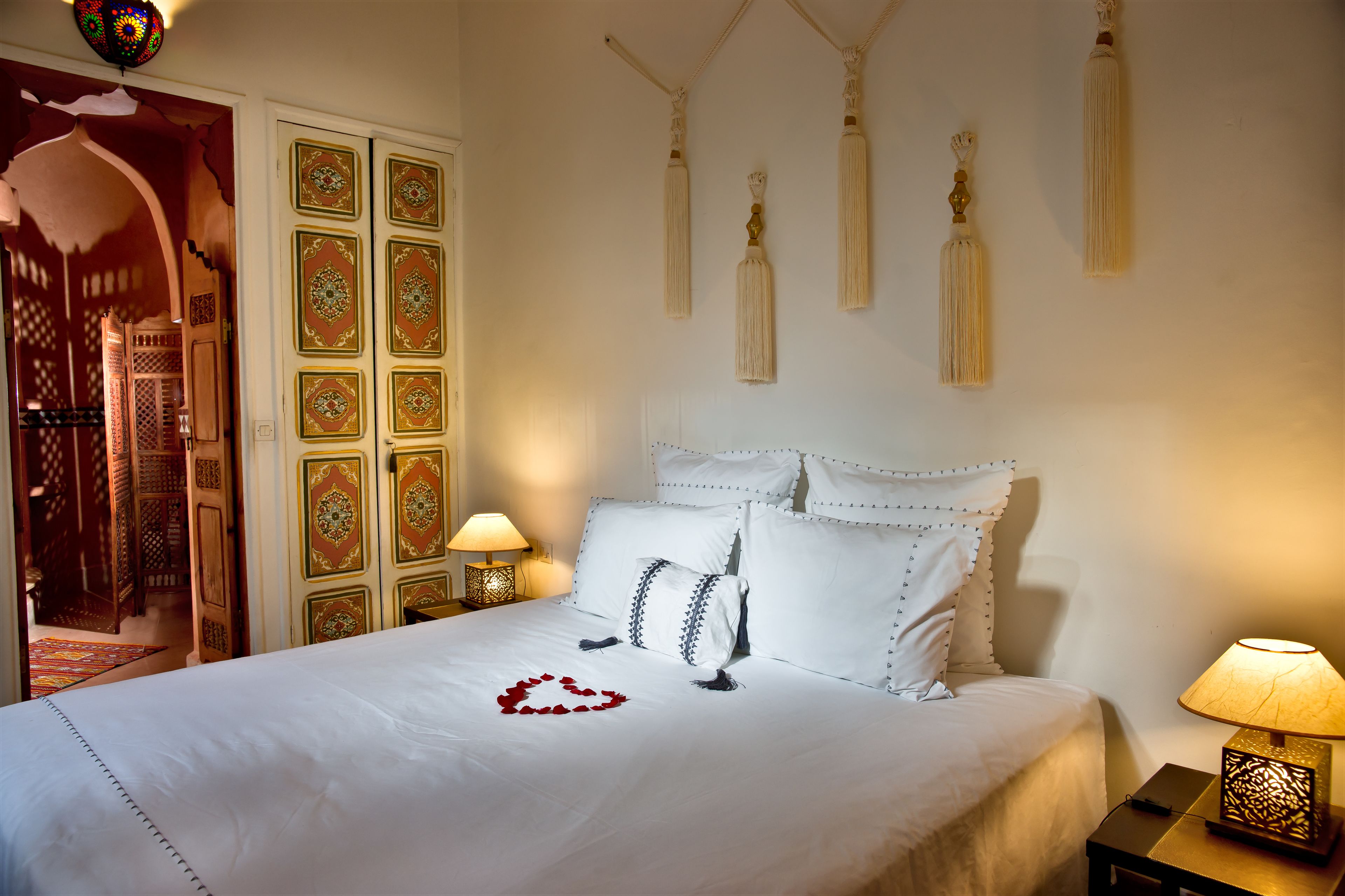 A neatly made bed with white linens and decorative pillows, a bedside lamp, and a wall with hanging tassels.