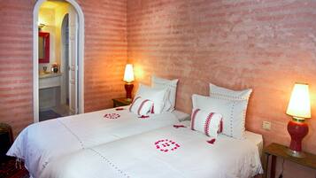 Una cama doble con sábanas blancas y almohadones decorativos, una mesita de noche con lámpara, y un espejo sobre un tocador con marco rojo.
