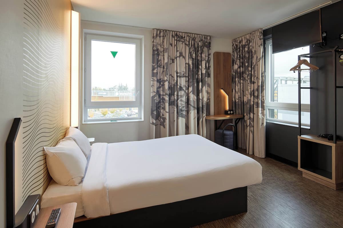 Ein Hotelzimmer mit einem Bett, einem Nachttisch mit Fernbedienung, einer Lampe und Blick auf die Stadt durch das Fenster.