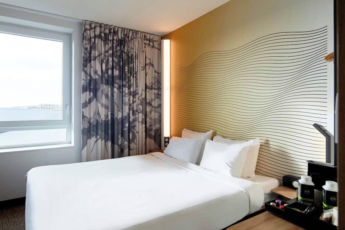 Ein Hotelzimmer mit großem Bett, Blick auf die Stadt, modernem Wanddesign und einem Nachttisch mit Kaffee und Snacks.