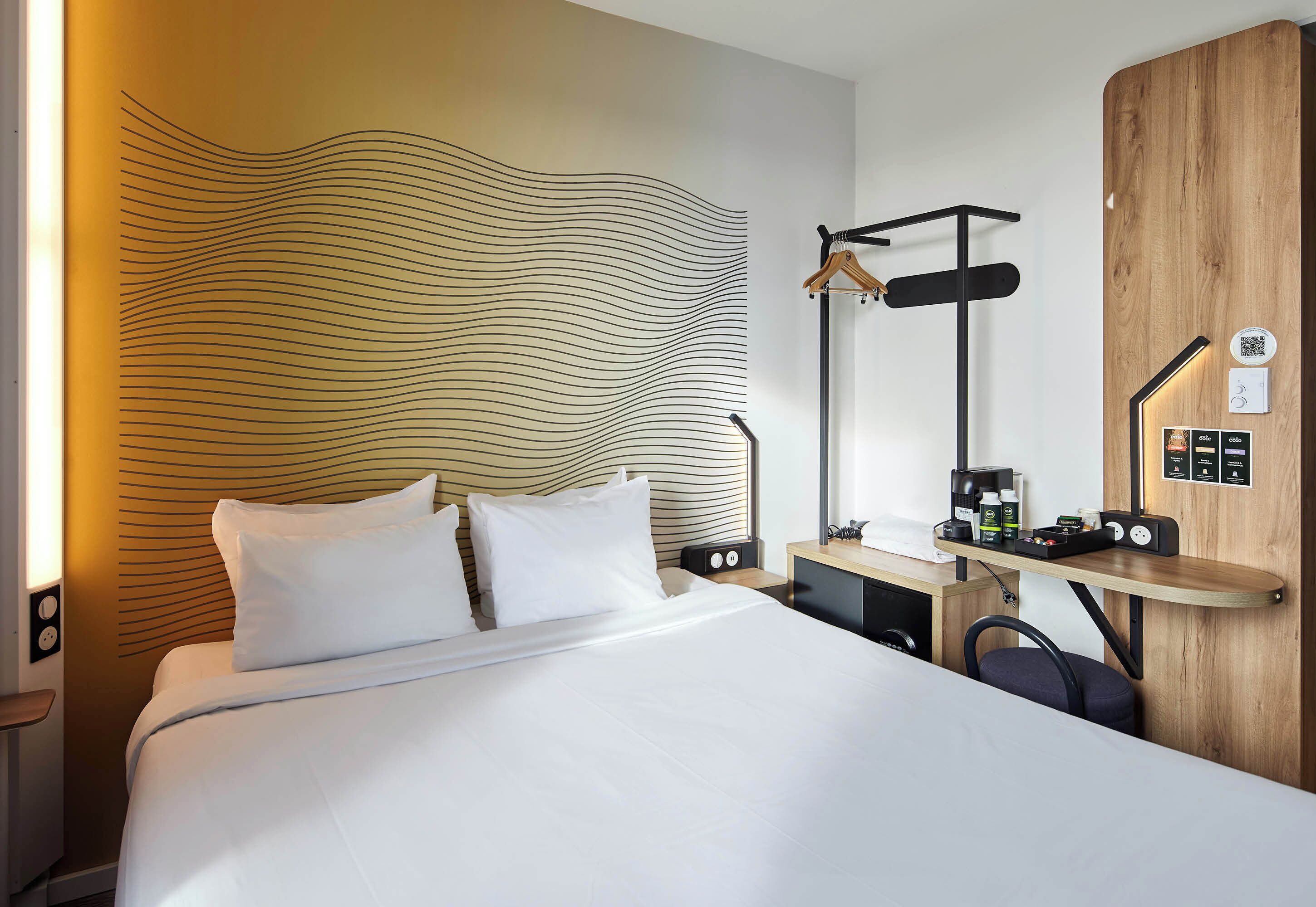 A modern hotel room with a large bed, a desk with a lamp, and a wooden panel wall.
