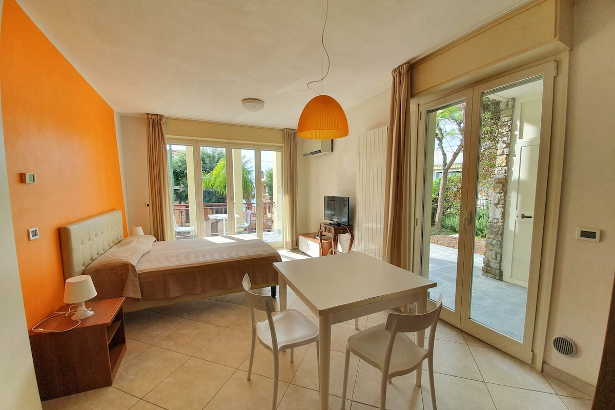 A hotel room with a bed, a desk with chairs, a television, and large sliding glass doors leading to an outdoor area.