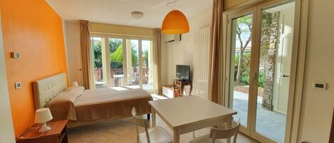 A hotel room with a bed, a desk with chairs, a television, and large sliding glass doors leading to an outdoor area.