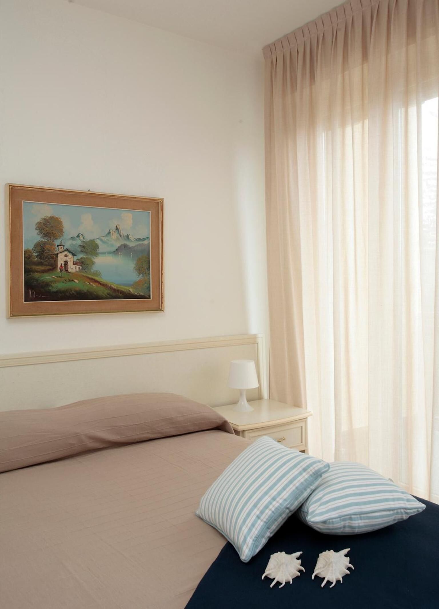 A bedroom with a bed, pillows, a nightstand, a lamp, a painting, and a window with curtains.