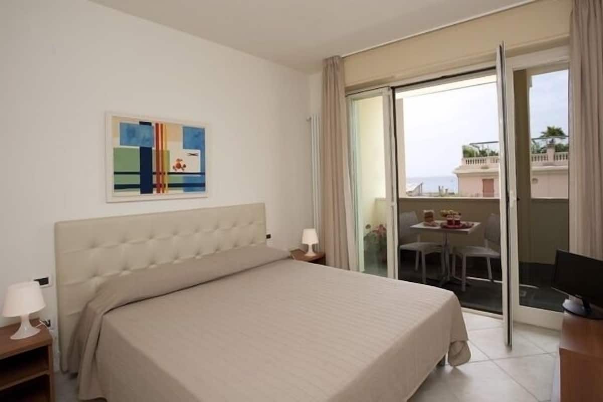 A hotel room with a bed, a nightstand, a lamp, a painting, and a balcony with a table and chairs.