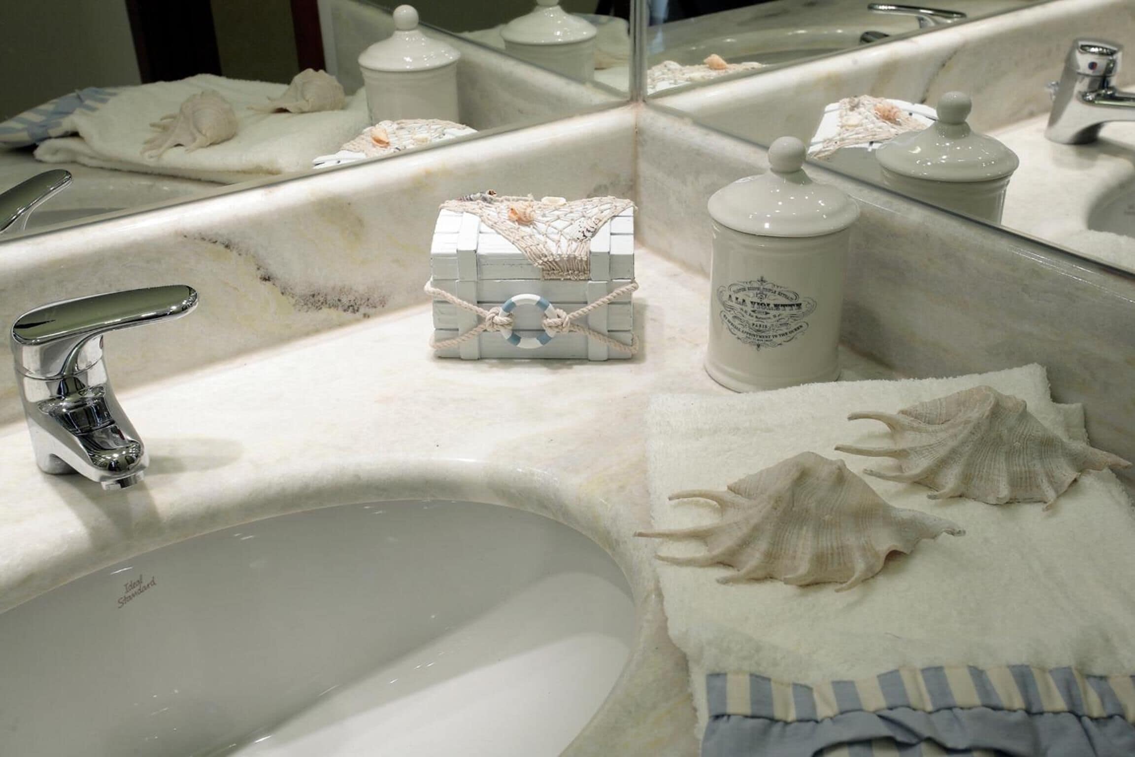 Un baño con un lavabo blanco, un espejo y un perchero para toallas. Hay conchas marinas y una pequeña caja decorativa sobre el mostrador.