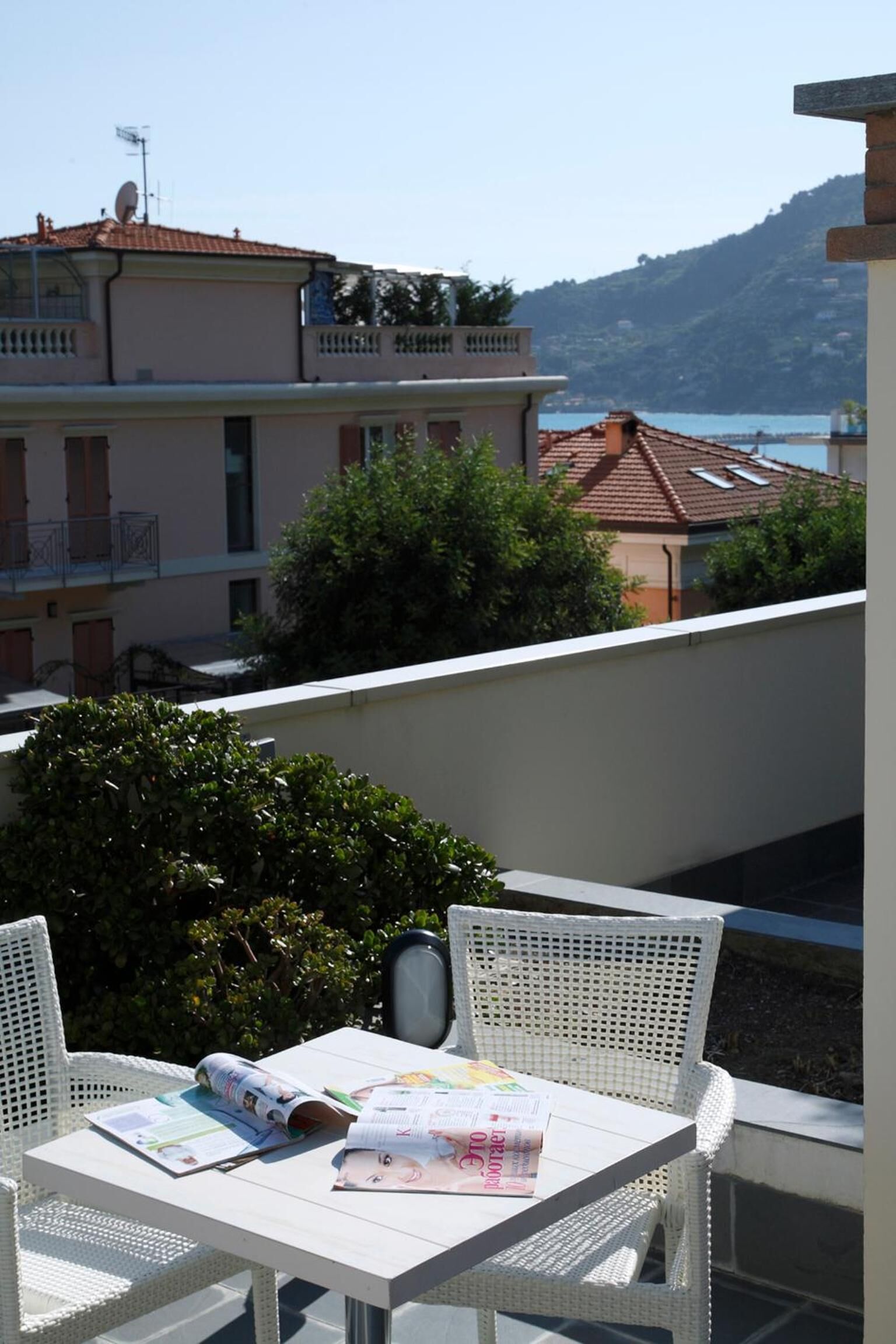 A balcony with white wicker furniture, a table with magazines, and a view of buildings and a body of water in the background.