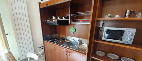 Cocina compacta con armarios de madera, un lavamanos de acero inoxidable, un microondas y una estufa a gas.