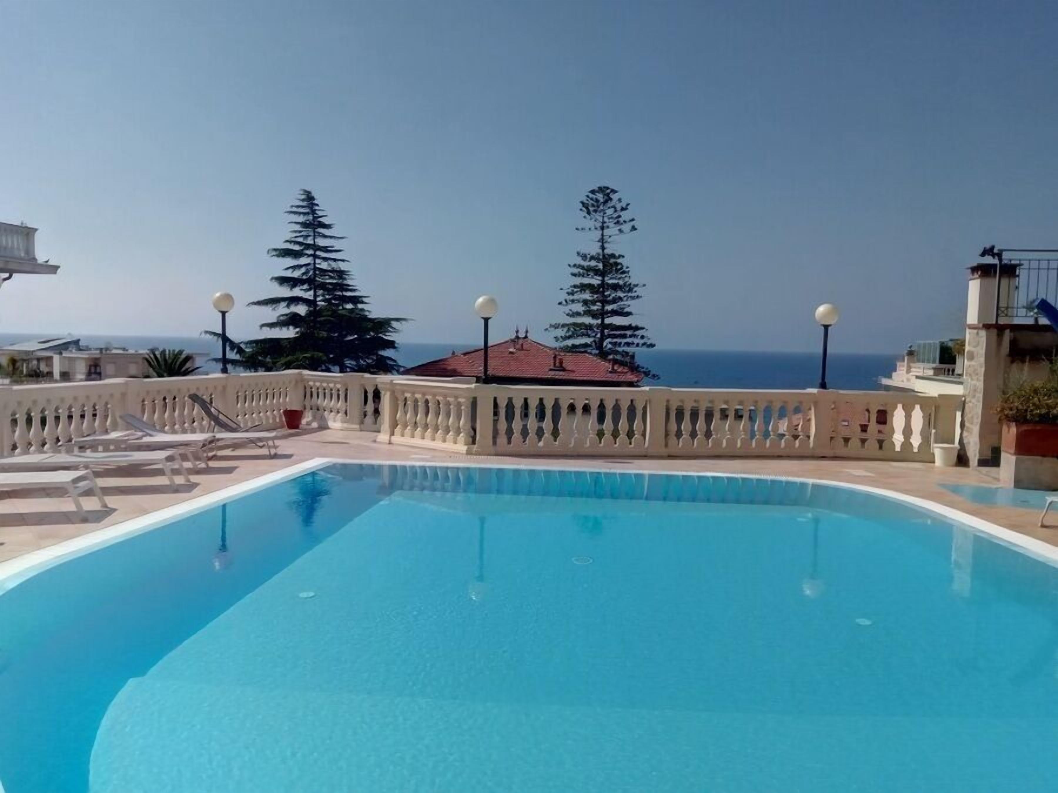 A swimming pool with a view of the sea, surrounded by a white balustrade and trees.