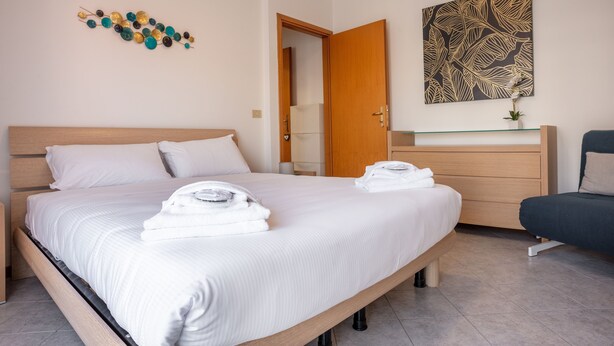 A modern hotel room with a large bed, a wooden headboard, a bedside table with a lamp, a small desk with a chair, and a decorative wall piece. - Residence Cava Zuccherina (Jesolo)