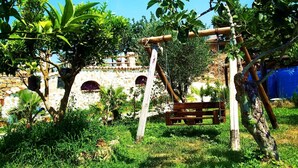 Un conjunto rústico de columpios de madera en un exuberante jardín, con un muro de piedra y un toldo azul al fondo.