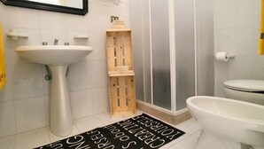 A bathroom with a white sink, toilet, and shower. There is a wooden crate and a black mat with white text. - Villetta Gardenia (Morciano di Leuca)