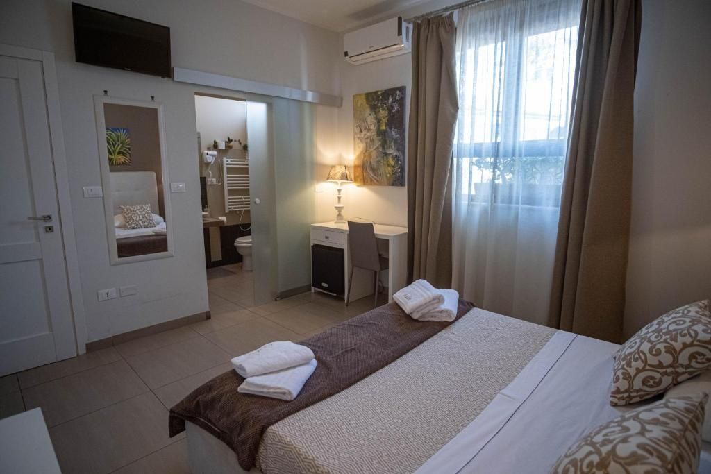Une chambre d’hôtel avec un lit, un bureau, une chaise, une fenêtre avec des rideaux et une salle de bain visible à travers une porte ouverte.