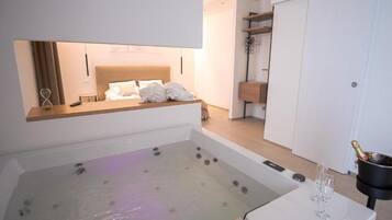 A modern hotel room with a large bathtub, a bed with white linens, a minibar, and a closet.