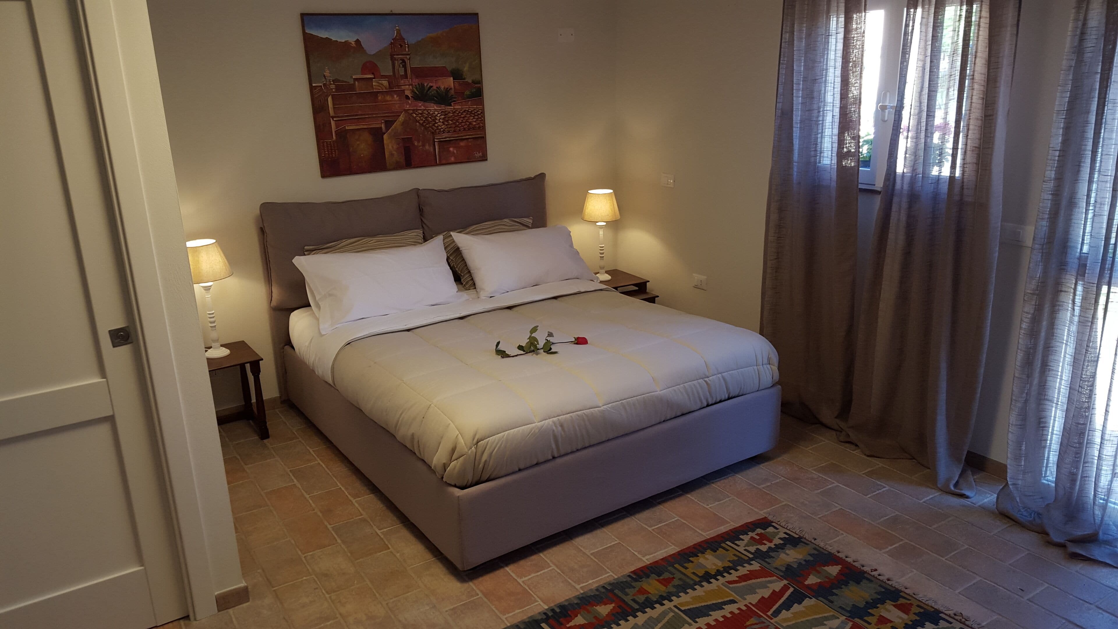 A bedroom with a bed, two bedside lamps, a painting on the wall, and a patterned rug.