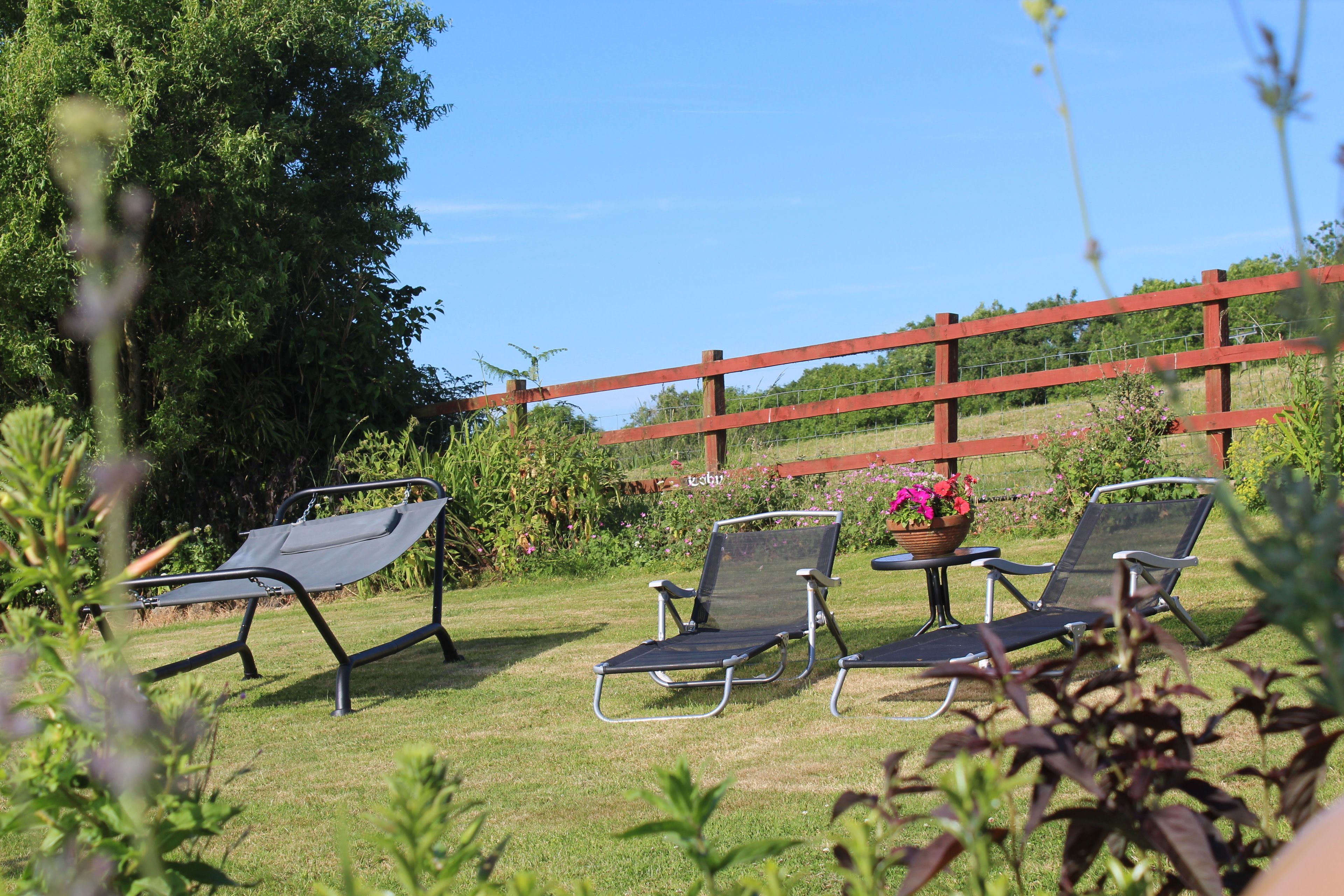 A garden with two lounge chairs and a table, surrounded by greenery and a red fence.