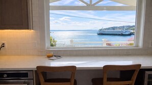A kitchen with a large window overlooking a body of water and a ferry. There are two wooden chairs at the counter. - Front Row Waterfront Views— Steps to Beach, Town & Ferry (Kingston)