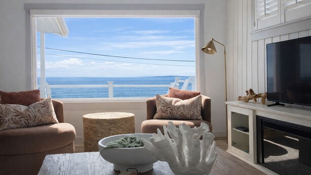 A living room with a large window overlooking the sea, a fireplace, and a television on a mantel. - Front Row Waterfront Views— Steps to Beach, Town & Ferry (Kingston)