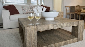 A rustic wooden coffee table with two wine glasses and a white bowl, placed in a living room with a white sofa and floral cushions. - Front Row Waterfront Views— Steps to Beach, Town & Ferry (Kingston)