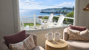A balcony with a view of a cruise ship, white outdoor furniture, and a wicker side table with two white mugs. - Front Row Waterfront Views— Steps to Beach, Town & Ferry (Kingston)