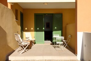 A small outdoor patio with a table and chairs, a folding chair, and a door with green shutters.