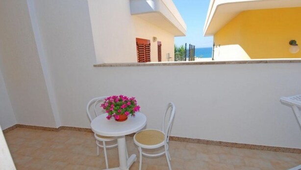 A small outdoor patio with a white table and two chairs, a potted plant with pink flowers, and a yellow building in the background. - Villetta Iris (Morciano di Leuca)