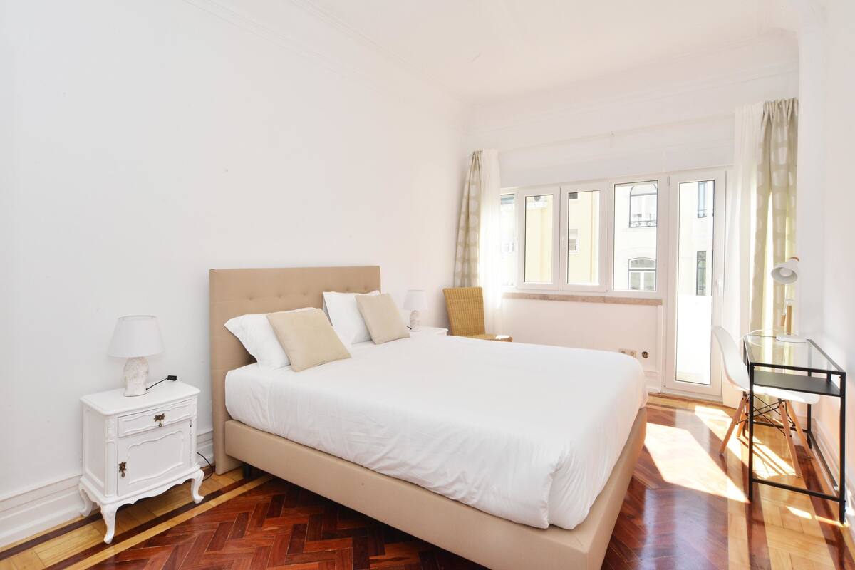 A bedroom with a large bed, a nightstand, a chair, and a window with curtains.