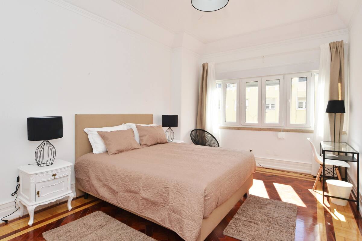 A bedroom with a large bed, two bedside tables with lamps, a chair, a desk with a lamp, and a window with curtains.